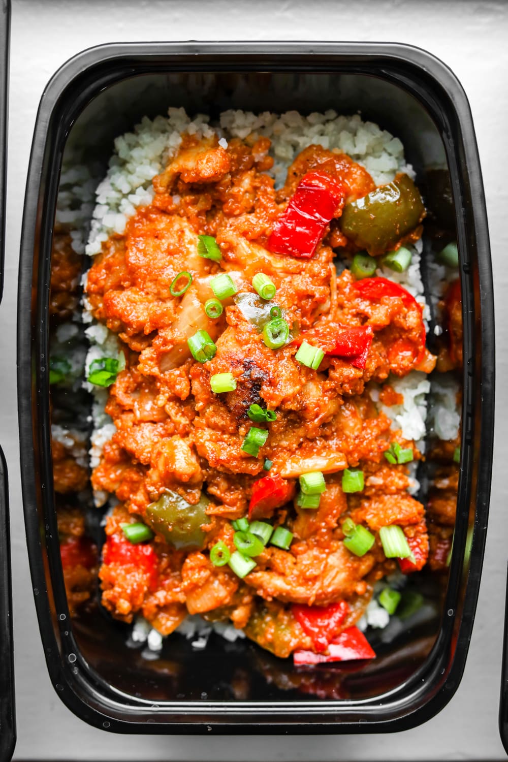 A meal prep container filled with cauliflower rice, BBQ chicken, yellow onion, and bell peppers.