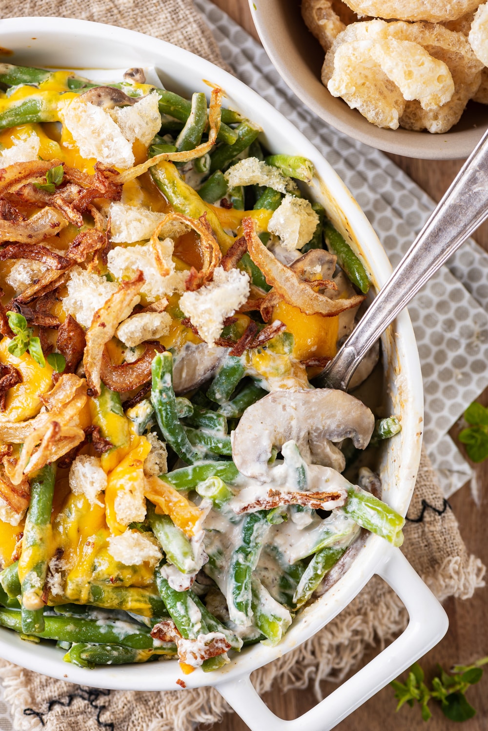 A mixture of green beans, creamy mushrooms, melted cheese, crispy onions, and pork rinds in a white casserole dish, and a fork placed in the dish as well.