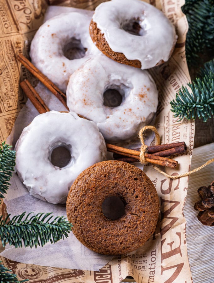 Four glazed gingerbread donuts and one plain gingerbread donut scattered on top of a piece of paper. Two sticks of cinnamon are surrounding the donuts on each side. Pine branches are at the bottom left and top right of the paper.