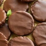 Chocolate coated cookies stacked on top of one another on a piece of brown parchment paper with a mint leaf next to them.