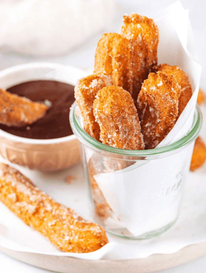 Glass cup filled with six churros. The glass cup is on a piece of white parchment paper on a plate and there is one churro next to the cup on the parchment paper and a cup of chocolate sauce behind it.
