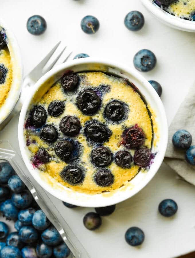 A blueberry muffin in a white mug. There are loose blueberries all around the mug.