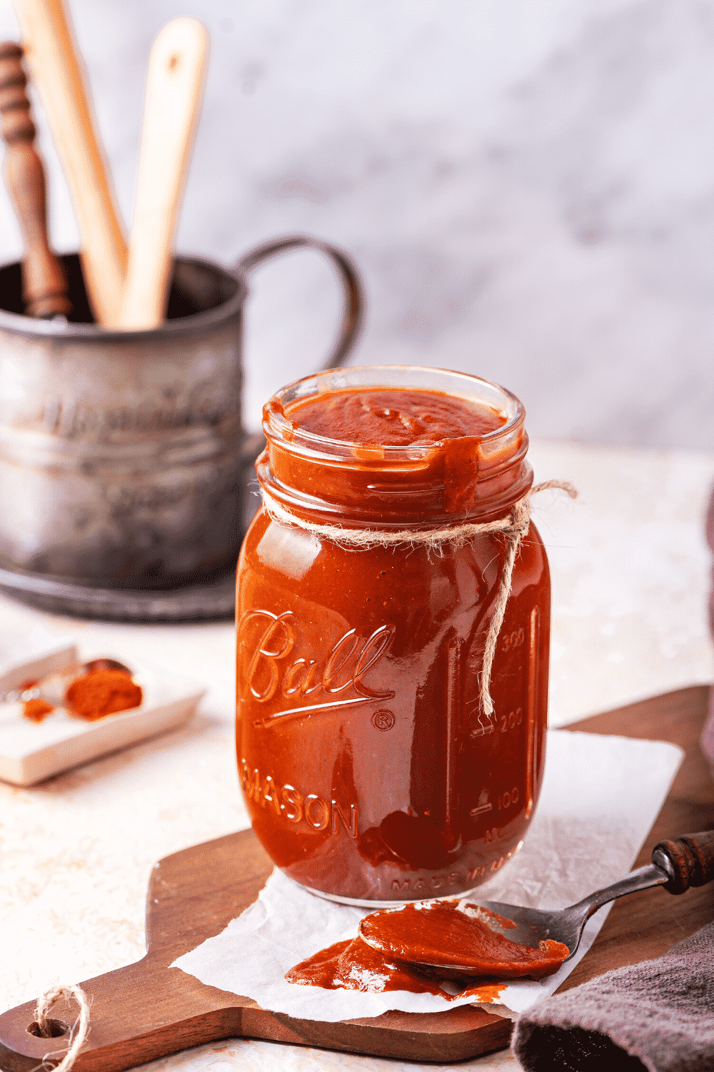 A glass jar filled with barbecue sauce. The jar is on a paper towel on a wooden cutting board and there is a spoon with barbecue sauce it on the paper towel.