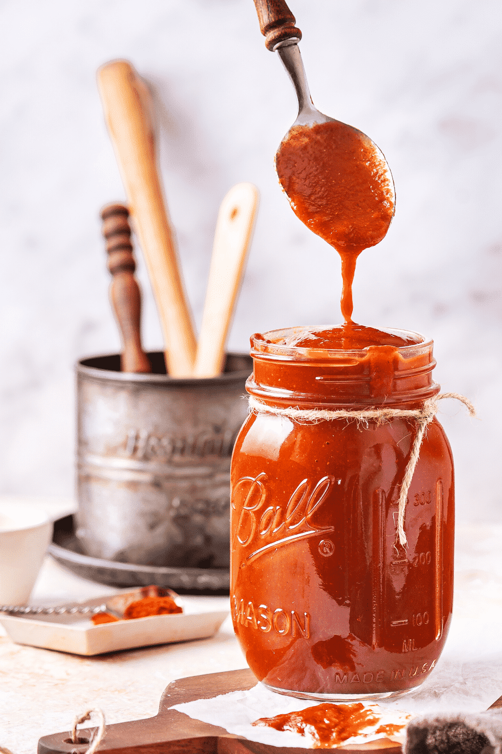 A glass jar filled to the top with BBQ sauce. A spoon is hovering over the jar dripping barbecue sauce down from it into the top of the jar.
