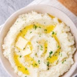 A white bowl filled with cauliflower mashed potatoes with two squares of butter and some melted butter on top. The balls on a white tablecloth on a gray counter and there is part of a wooden cutting board behind it.
