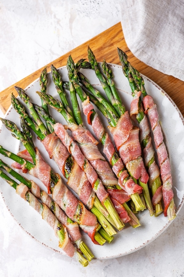 A white plate with asparagus wrapped in bacon on it. The plate is on a wooden cutting board that is sat on a white counter.