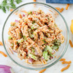 Tuna salad in a bowl. There is a stalk of celery, a red onion, a blue napkin, macaroni, and dill surrounding the bowl.