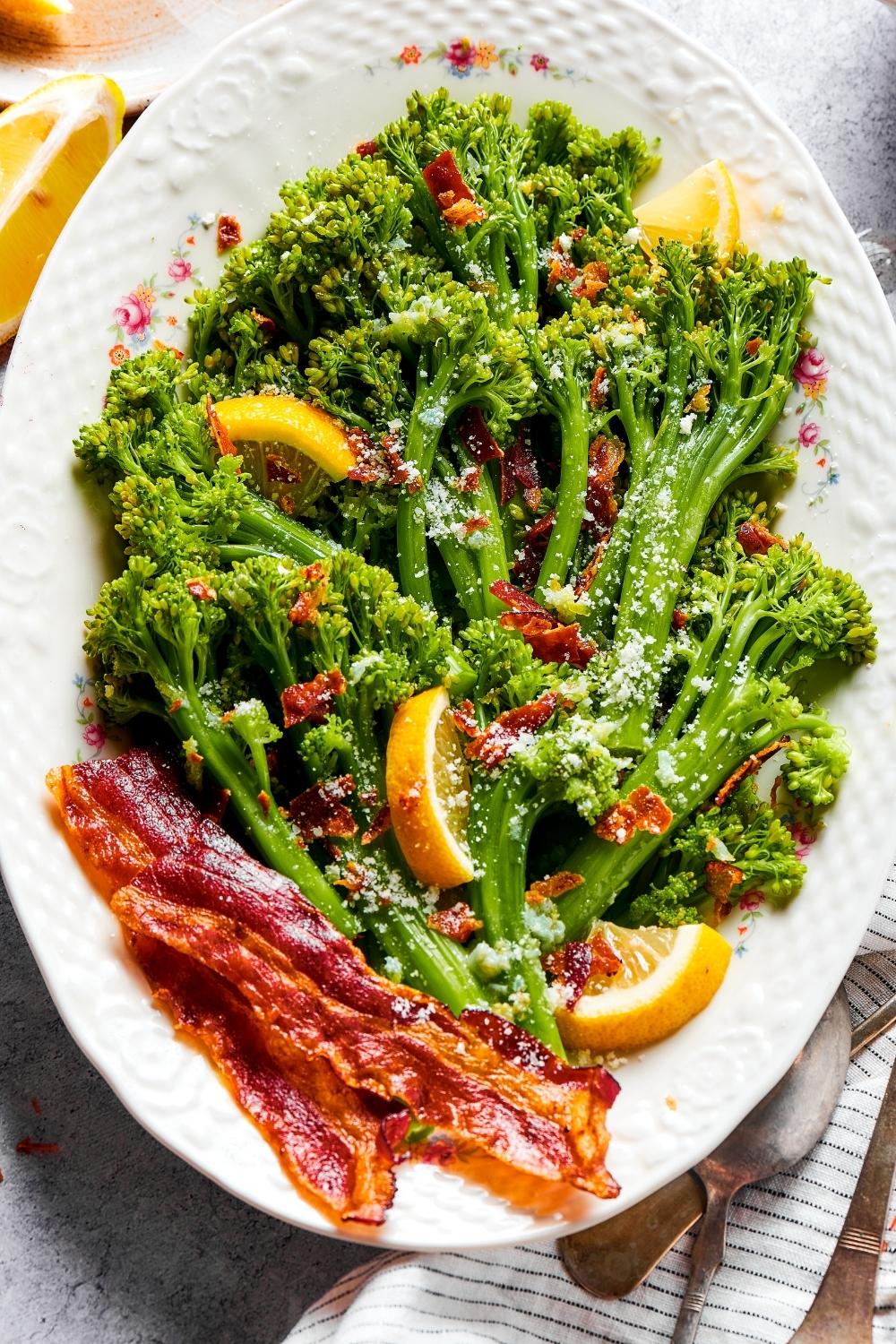 Pieces of bacon on top of broccolini on a plate with lemon wedges and a few slices of bacon.