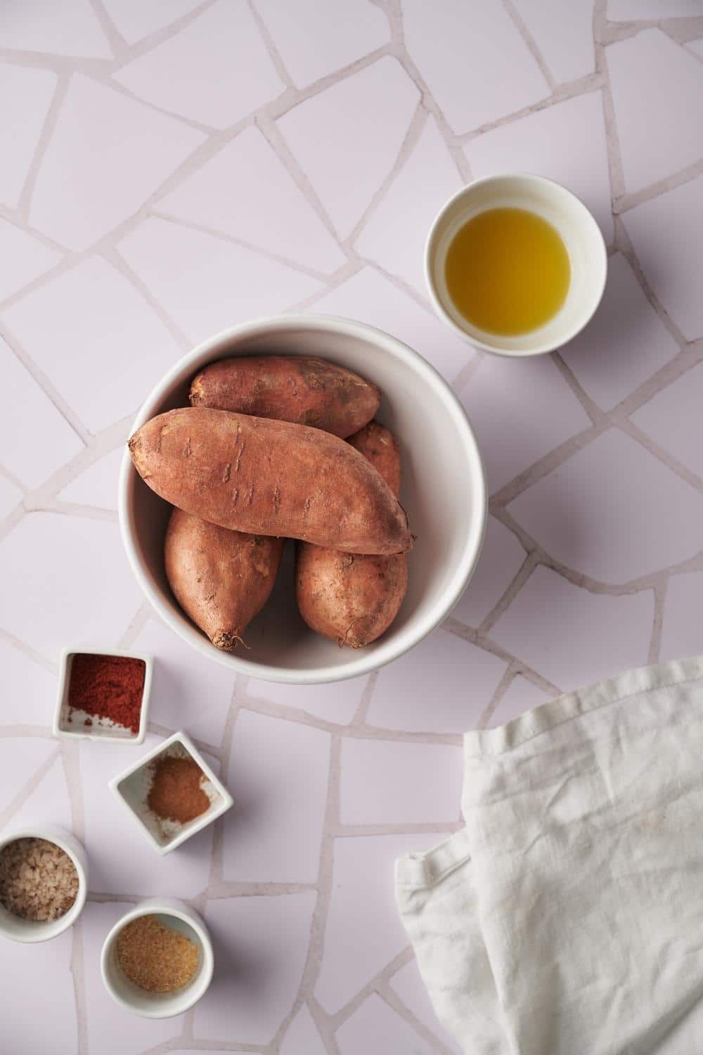 A large bowl filled with 4 sweet potatoes. Surrounding it is a small bowl of olive oil and smaller bowls filled with individual spices.