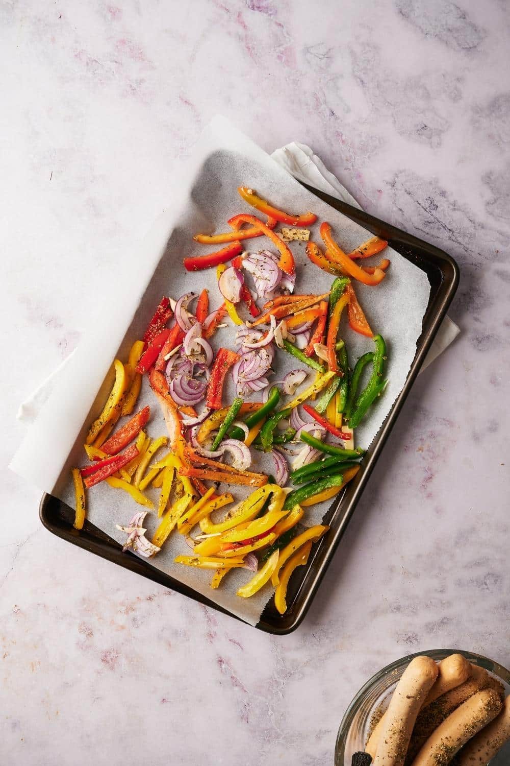 Sliced bell peppers and onions seasoned with herbs on a parchment paper lined baking tray.