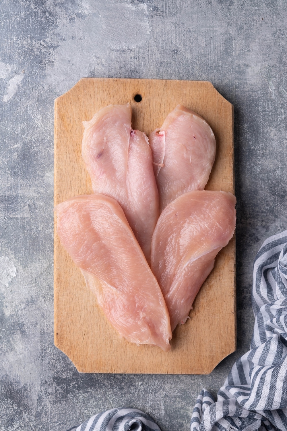 Two chicken breasts cut into 4 equally sized slices, fanned out on a wood cutting board. The board is on a grey countertop next to a blue and white striped tea towel.