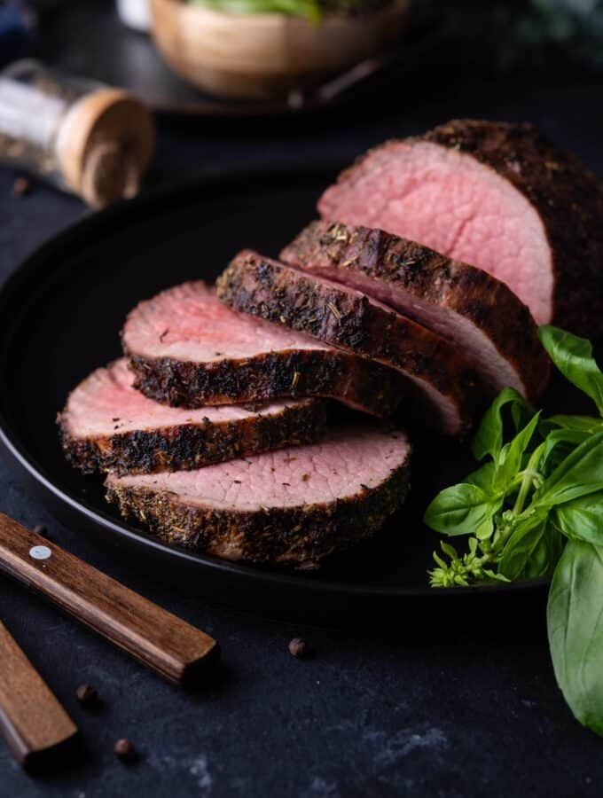 Medium rare grilled beef tenderloin, served and served on a black plate with basil leaves.