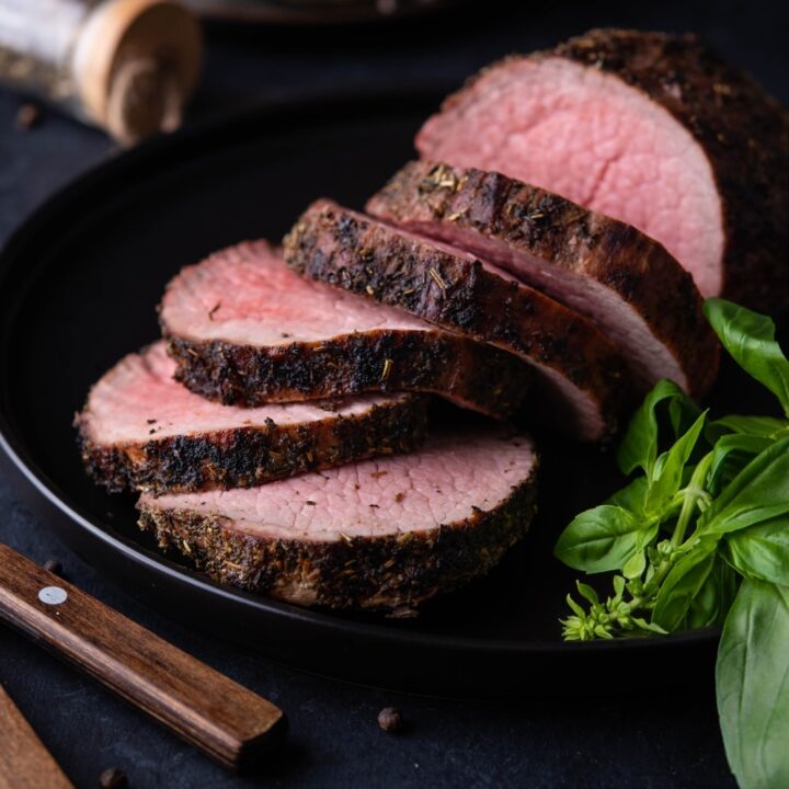 Medium rare grilled beef tenderloin, served and served on a black plate with basil leaves.