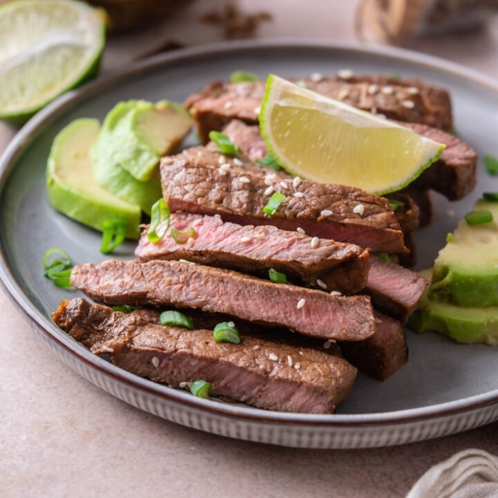 Sirloin tip steak on a plate, sliced and served with avocado slices and lime wedges, garnished with chopped green onions and sesame seeds.