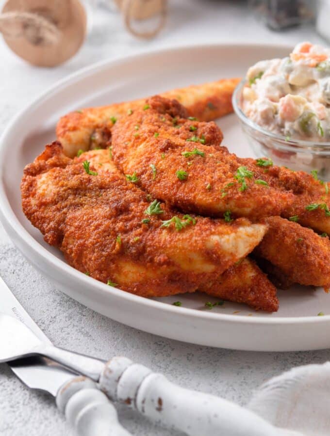 Baked chicken tenders garnished with chopped parsley with a small bowl of creamy potato salad.