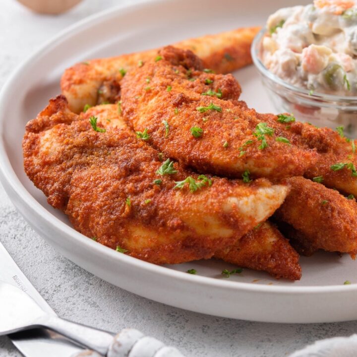 Baked chicken tenders garnished with chopped parsley with a small bowl of creamy potato salad.
