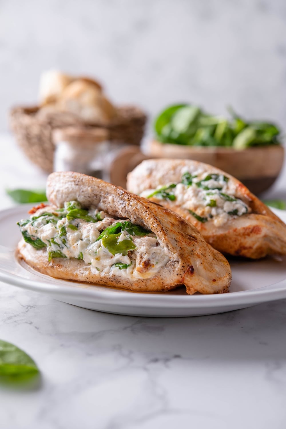 Two cream cheese and spinach stuffed chicke breasts on a plate, in front of bowls of bread and spinach.