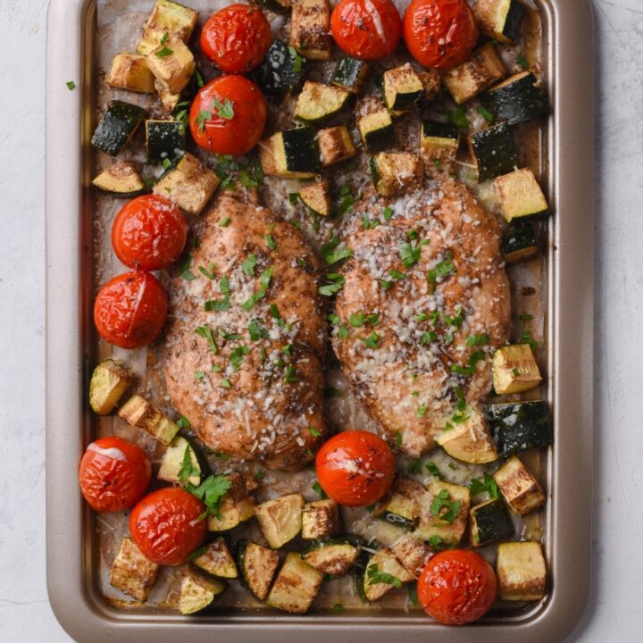 Seasoned baked chicken filets, cherry tomatoes, and zucchini on a sheet pan lined with parchment paper.