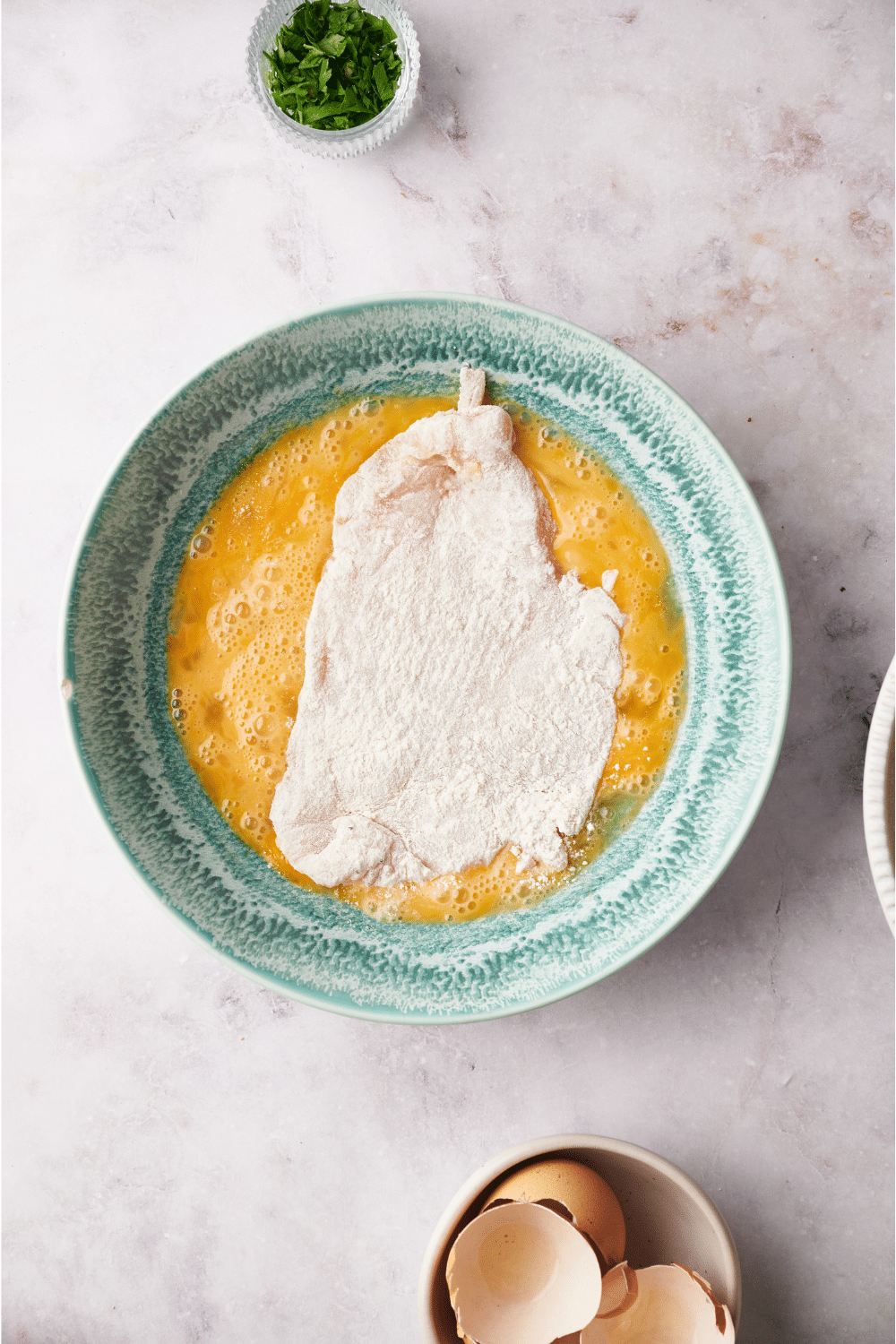 A blue bowl filled with a beaten egg mixture and a flour-coated chicken cutlet is in the bowl.