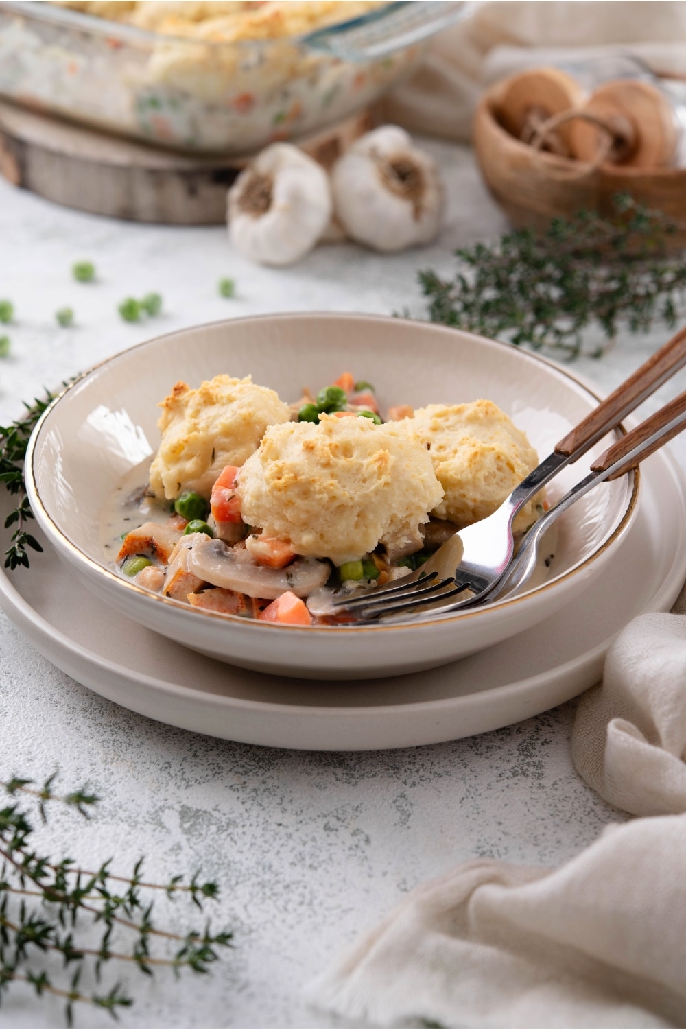 A dish of chicken and biscuits casserole in a white bowl on a white plate. A fork and spoon are resting in the dish. In the background there is a casserole dish, garlic cloves, and fresh herbs.