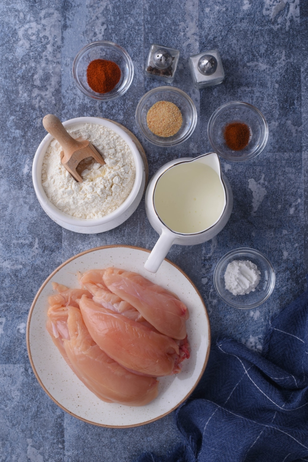 Paprika in a bowl, cayenne pepper in a bowl, garlic powder in a bowl, flour in a bowl, buttermilk in a pitcher, and chicken on a plate all on a grey counter.