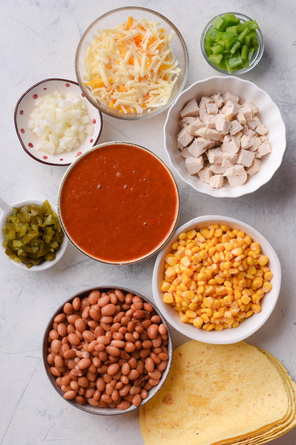An assortment of ingredients including bowls of enchilada sauce, diced chicken, pinto beans, corn, shredded cheese, diced green peppers, diced onion, green chiles, and a stack of tortillas all on a white counter.