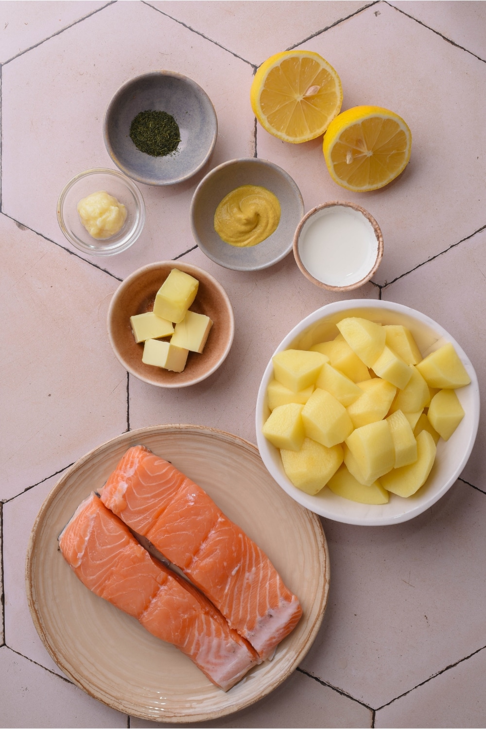 A plate with two fillets of salmon on it. A bowl of mashed potatoes. A cup of butter, a cup of mustard, a cup of dried dill, and a cup of garlic all on a white counter.