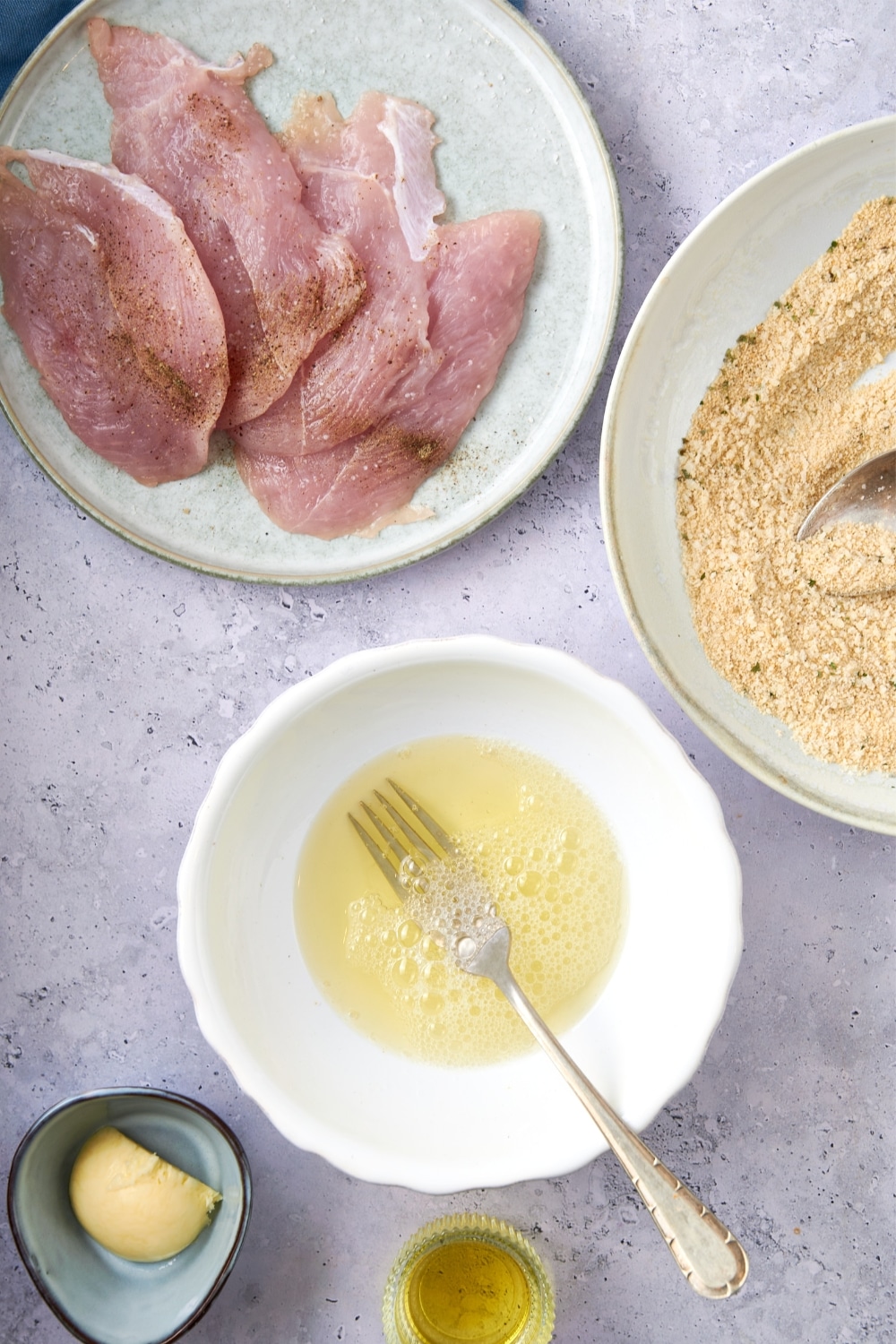 A plate of turkey breast cutlets next to shallow bowls of egg whites and seasoned breadcrumbs and smaller bowls of butter and olive oil.