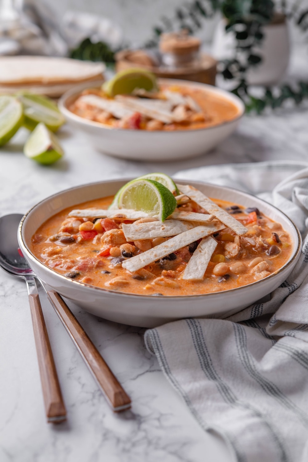 A bowl of creamy soup with diced peppers, corn, beans, and chicken, with crispy tortilla strips and lime wedges garnished on top.