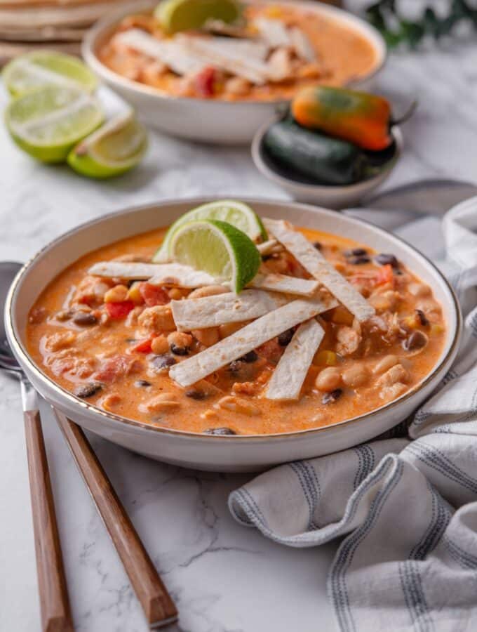 A bowl of creamy soup with diced tomatoes, corn, beans, and chicken, with crispy tortilla strips and lime wedges garnished on top.
