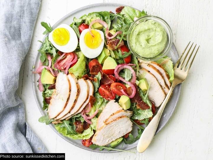 Sliced chicken breast, red onion, avocado, a sliced egg, and lettuce on a plate with a cup of green goddess dressing.