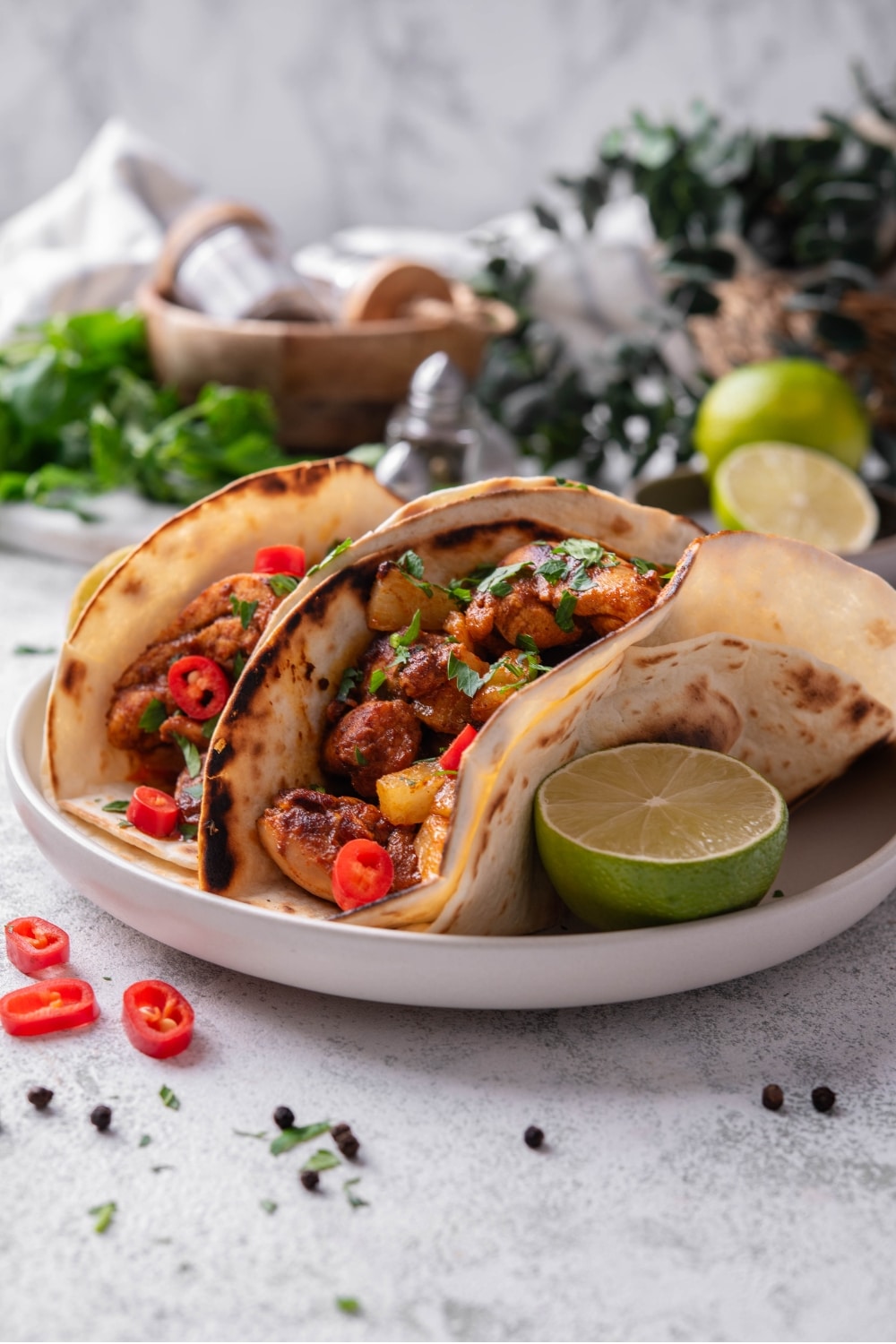 A plate with two chicken al pastor tacos with a halved lime on the side. The tacos contain chicken, pineapple, and peppers with cilantro on top. There is a wooden bowl with seasonings and limes in the background.