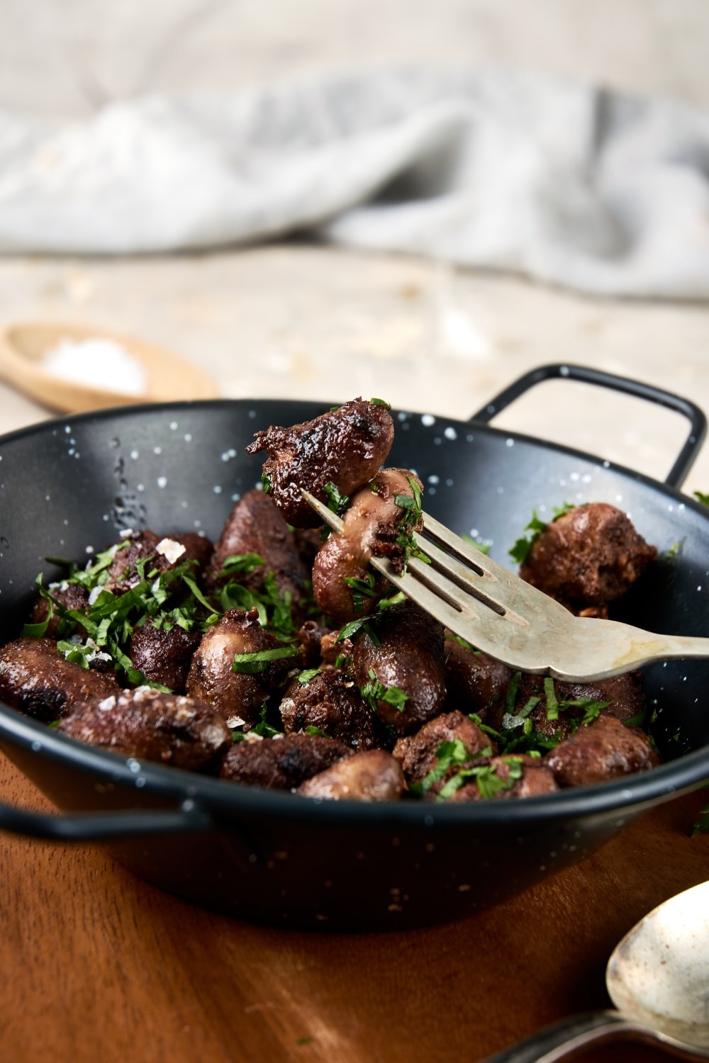 A ceramic bowl filled with cooked chicken hearts topped with fresh parsley. The bowl is on a wooden server and there is a fork taking a bite of chicken hearts.