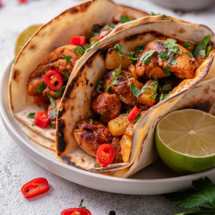 A plate with two chicken al pastor tacos with a halved lime on the side. The tacos contain chicken, pineapple, and peppers with cilantro on top. There is a bowl of limes in the background.