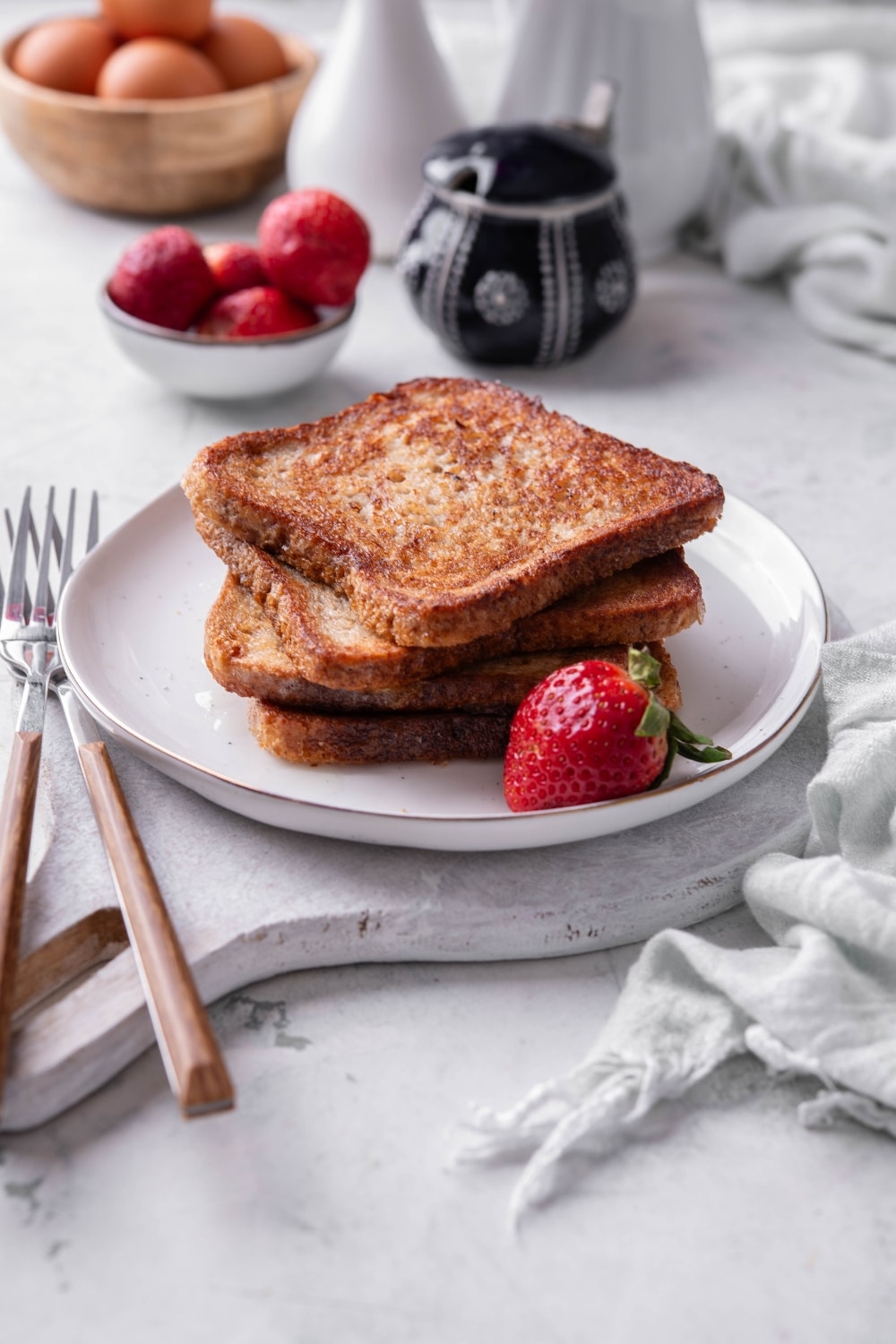 Four slices of french toast stacked on a white plate on top of a serving board. There is a strawberry and forks on the side.