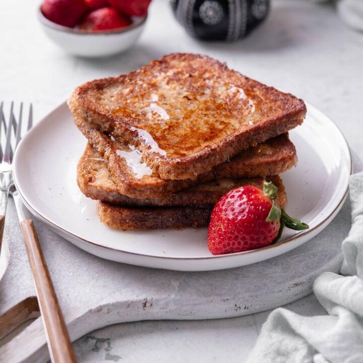 Four slices of french toast with syrup stacked on a white plate on top of a serving board. There is a strawberry and fork on the side.