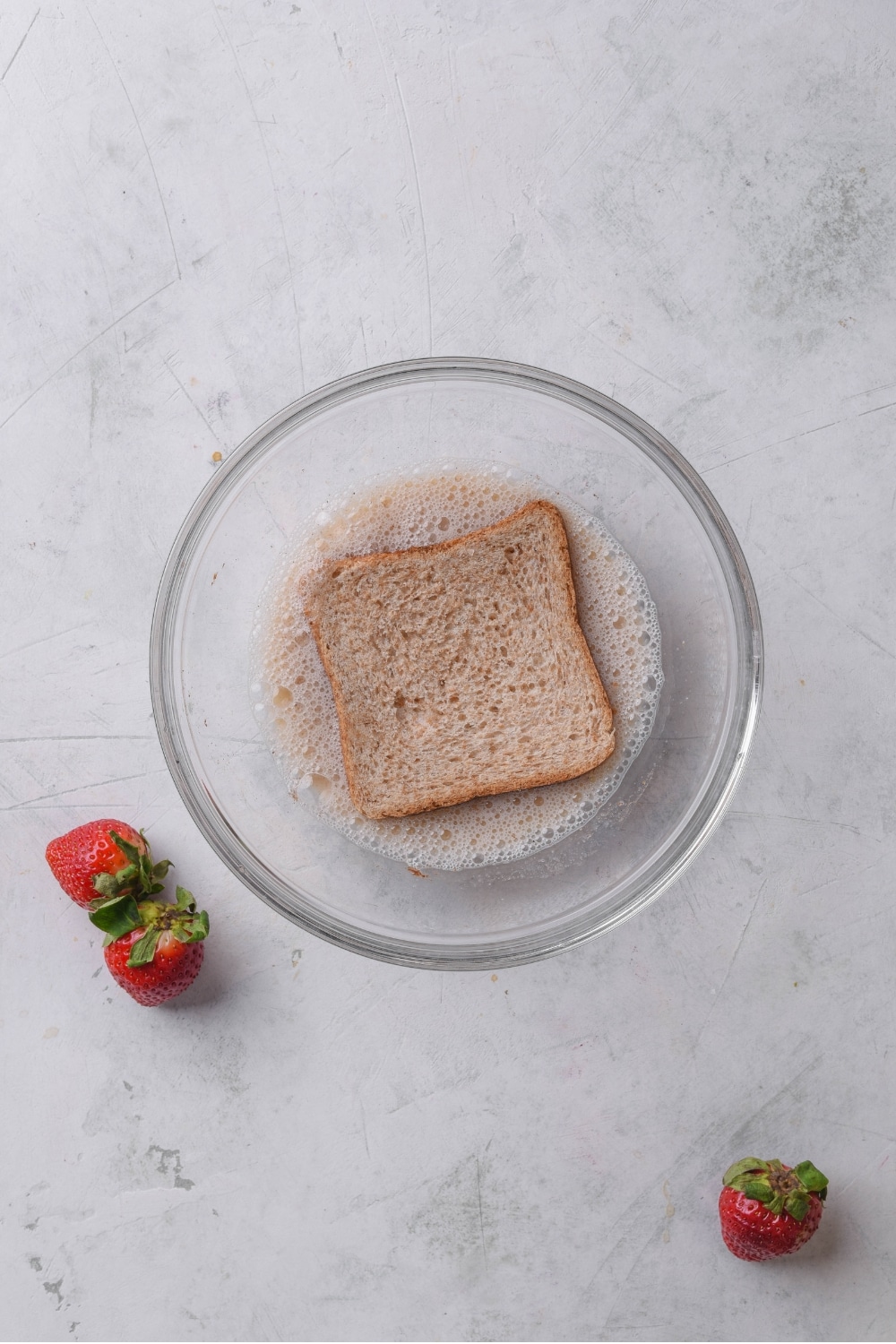 An overhead shot of a glass bowl with a slice of bread soaking in an egg and milk mixture.