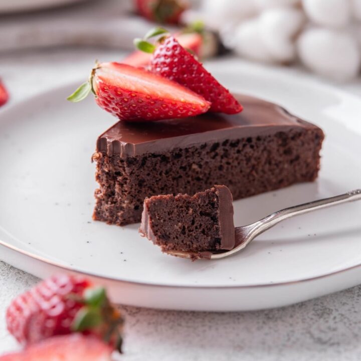 A slice of chocolate protein cake topped with sliced strawberries on a white ceramic plate. There is a fork with a bite of the cake on the plate.