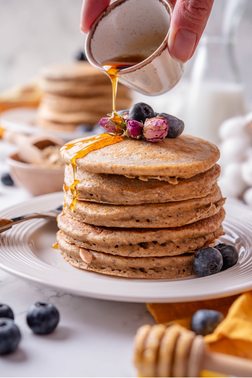 A stack of five high fiber pancakes on a white ceramic plate. The pancakes are topped with blueberries and dried flower buds and there is a hand pouring maple syrup from a ceramic ramekin over the pancakes.