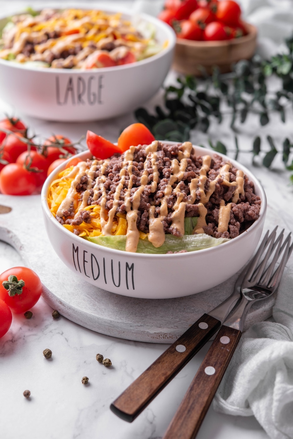 A bowl of Big Mac salad with shredded lettuce, shredded cheddar cheese, and ground beef drizzled with Big Mac salad dressing on top. The bowl has the word "Medium" written on it, and is served on a ceramic platter. There is another Big Mac salad bowl in the background with the word "large" written on it.