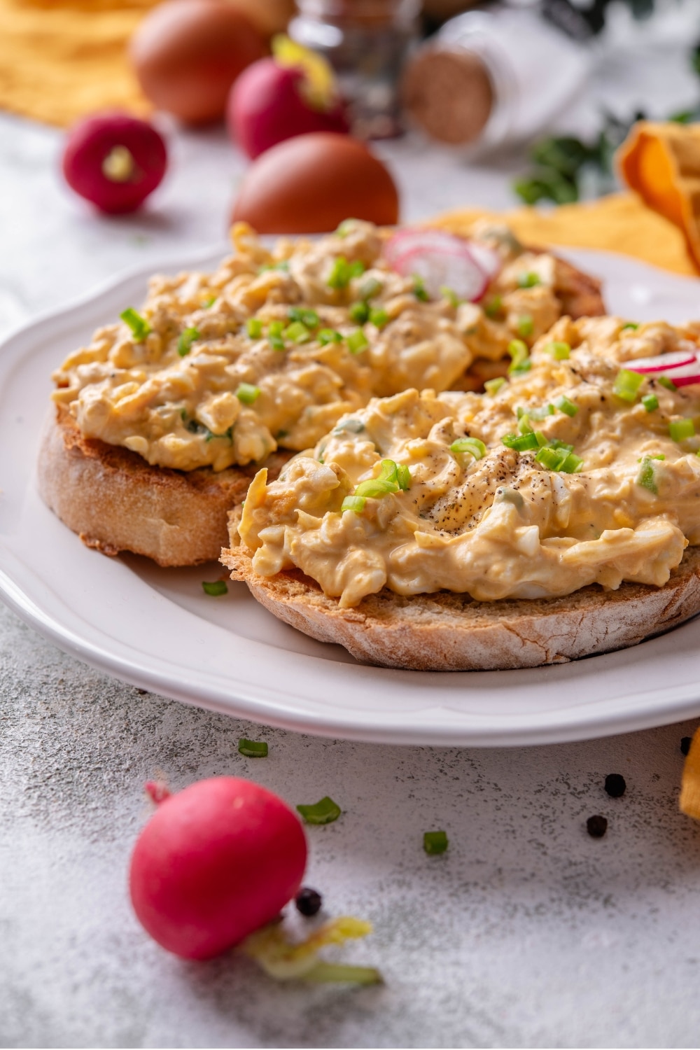 A plate of two slices of toasted bread with healthy egg salad on top. The egg salad is garnished with green onion and sliced radishes, with whole radishes surrounding the plate.