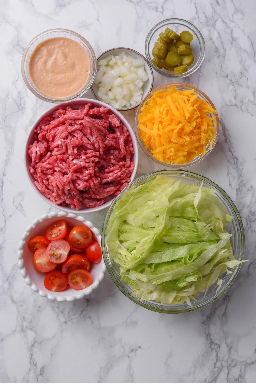 An overhead shot of several bowls of various sizes and shapes containing the ingredients for Big Mac salad including raw beef, shredded iceberg lettuce, shredded cheddar cheese, diced white onion, halved cherry tomatoes, chopped pickles, and Big Mac salad dressing.