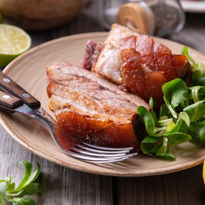 A tan ceramic plate with two pieces of sliced pork belly with a small side of greens. There are two forks on the plate and a halved lemon in front of the plate.