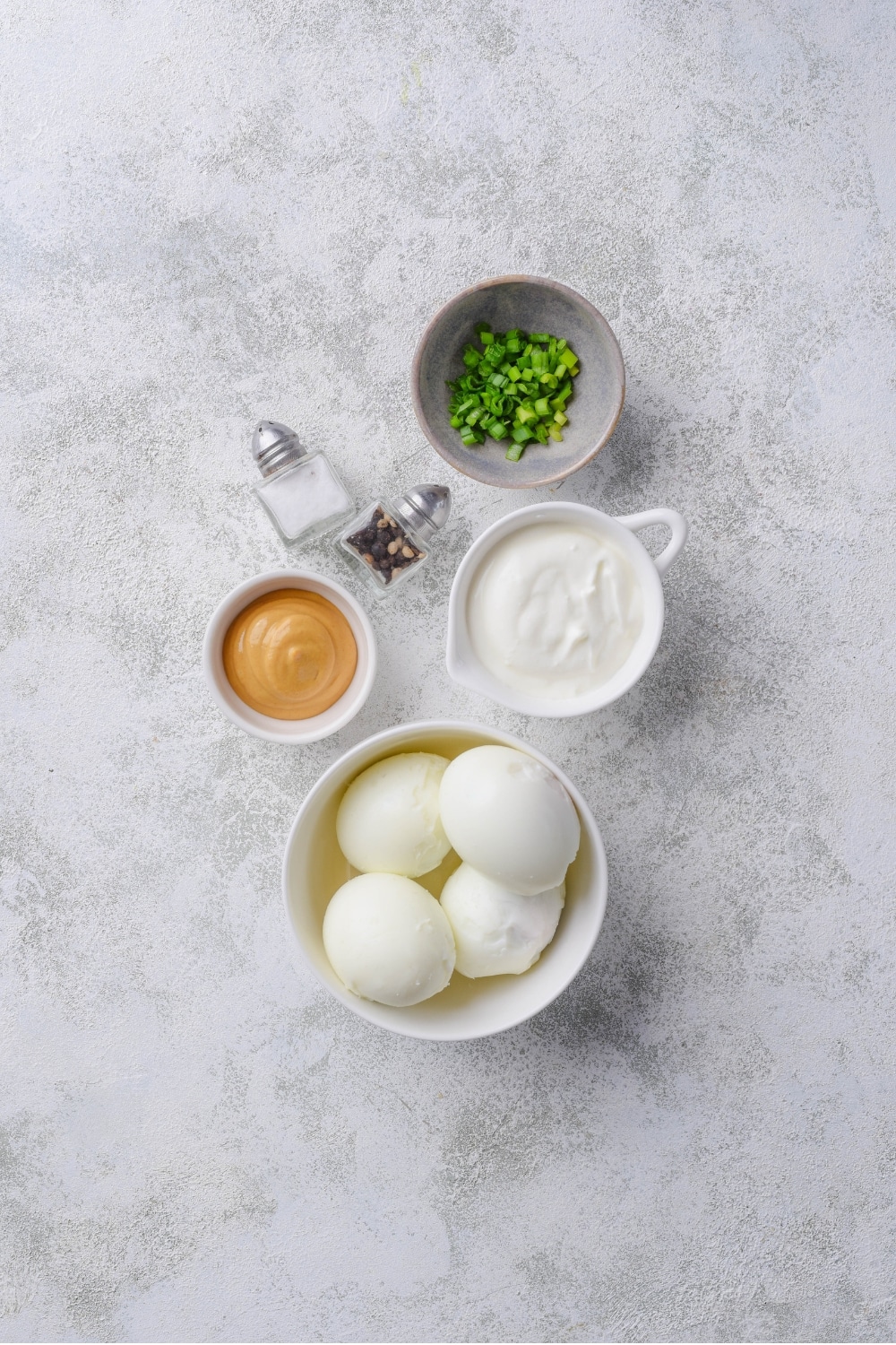 An overhead shot of several bowls filled with various ingredients for healthy egg salad including hard boiled eggs, yogurt, mustard, green onion, and salt and pepper.