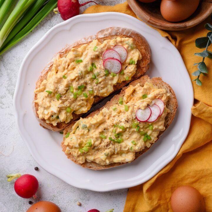 An overhead shot of a plate of two slices of toasted bread with healthy egg salad on top. The egg salad is garnished with green onion and sliced radishes, with whole radishes surrounding the plate. There is a small bowl of eggs next to the plate.