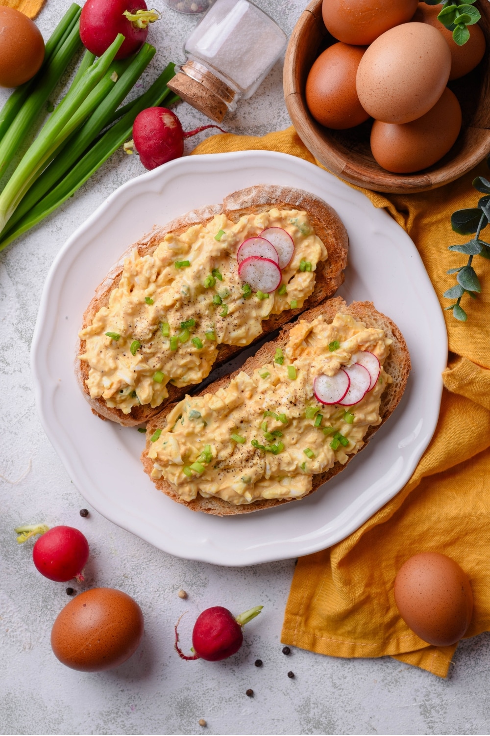 An overhead shot of a plate of two slices of toasted bread with healthy egg salad on top. The egg salad is garnished with green onion and sliced radishes, with whole radishes surrounding the plate. There is a small bowl of eggs next to the plate.