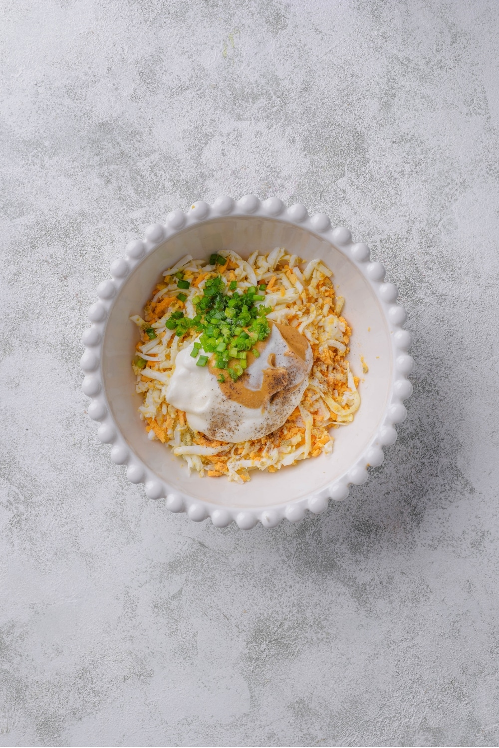 An overhead shot of a white porcelain bowl filled with grated hard boiled egg, greek yogurt, mustard, and chopped green onion.