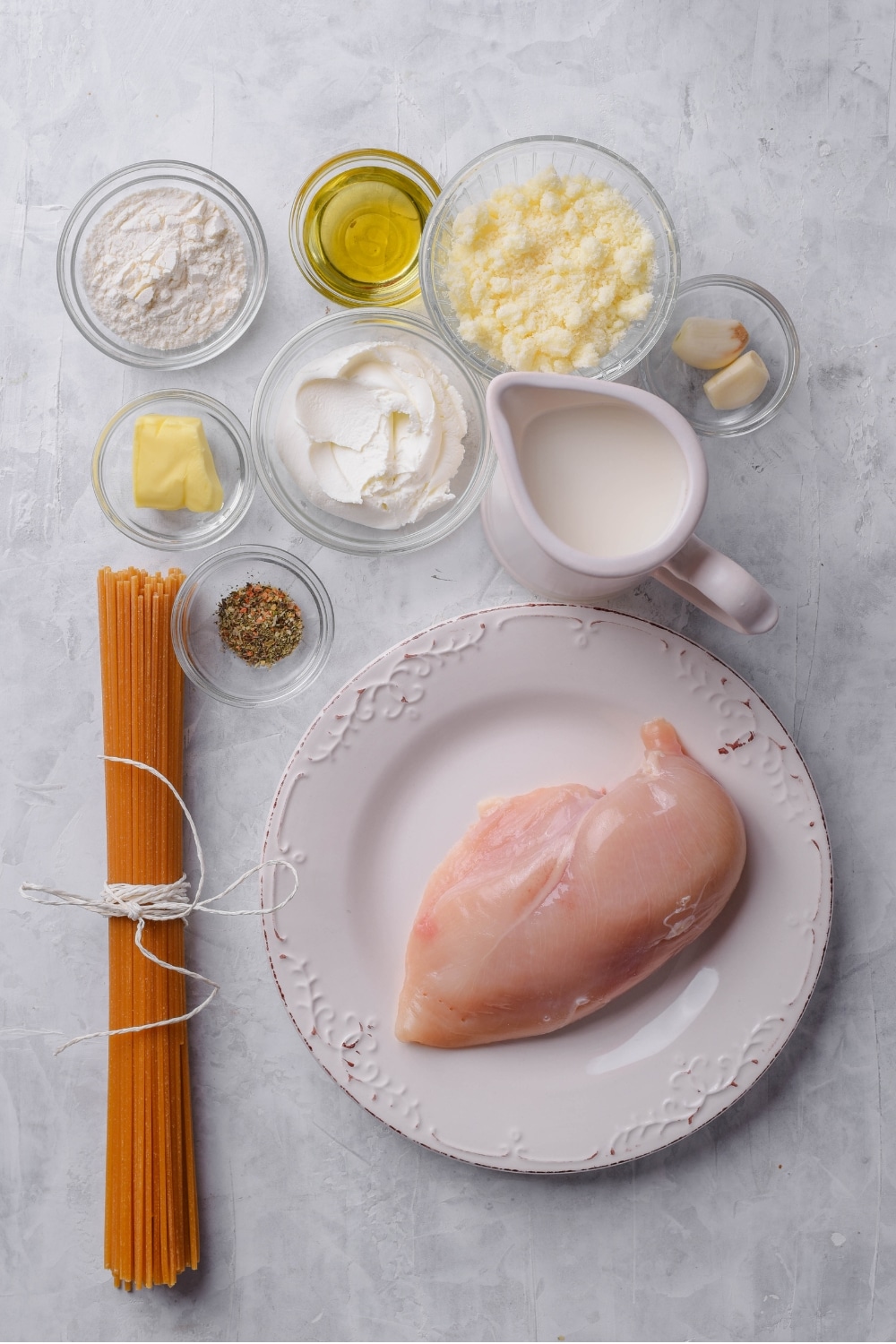 An overhead shot of several bowls of various shapes and sizes containing the ingredients for low calorie alfredo sauce including a plate of raw chicken breast, cream cheese, milk, garlic, butter, flour, seasonings, and spaghetti wrapped in string.