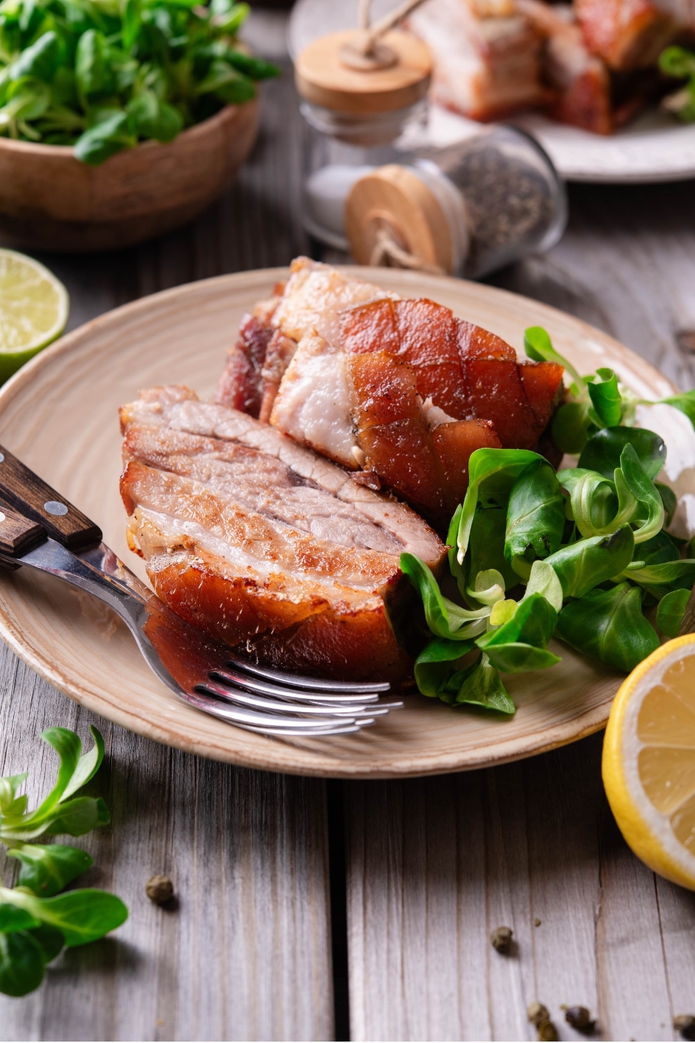 A tan ceramic plate with two pieces of sliced pork belly with a small side of greens. There are two forks on the plate and a halved lemon in front of the plate.