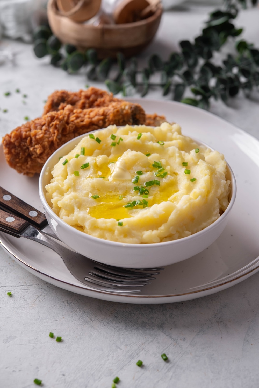A white ceramic bowl filled with gold mashed potatoes with chives and melted butter on top. The bowl is on a ceramic plate with a piece of friend chicken next to the bowl. There are two forks tucked next to the bowl.